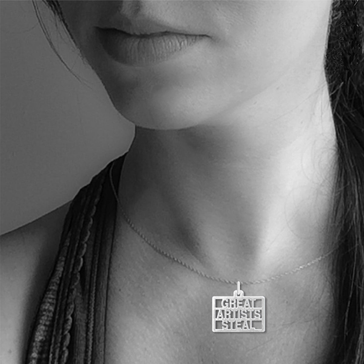 Great Artists Steal necklace worn on a silver chain, featuring a square silver pendant with cutout lettering displayed against the neckline of a model.