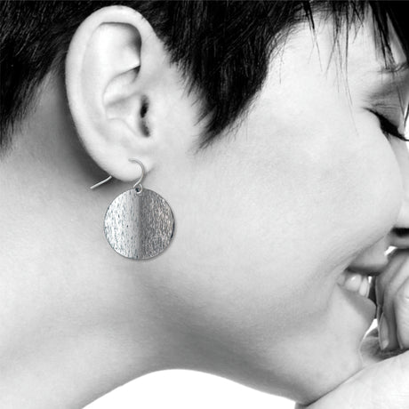 An Orb circular drop earring with a split-tone brushed silver finish, shown worn on ear with a sleek minimalist design inspired by Ellsworth Kelly’s sculptural artwork