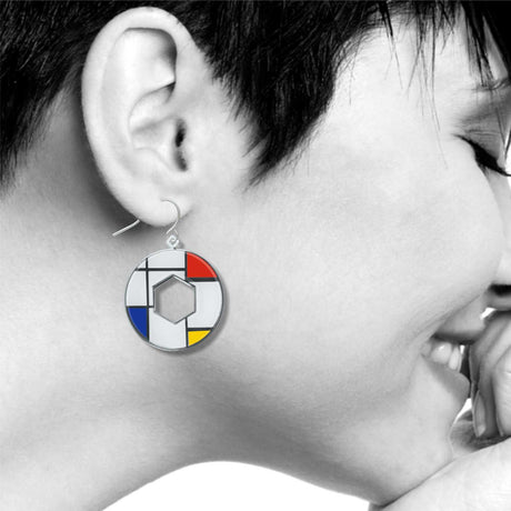 Model wearing a Utopia geometric dangle earring inspired by Mondrian artwork, featuring bold red, blue, yellow, black, and white enamel accents in a circular open-center design.