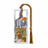 A silver metal bookmark featuring a design with a red-tailed hawk above Central Park, complete with a autumnal display of trees and a cityscape in the background. It includes a gold-colored tassel.